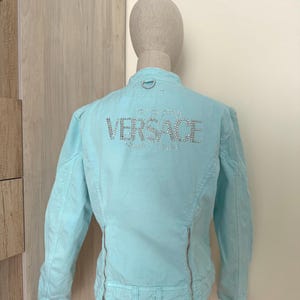 Może przedstawiać: Jasnoniebieska kurtka z napisem "VERSACE" wysadzanym srebrnymi kryształkami na plecach. Kurtka posiada zapięcie na zamek błyskawiczny i mały srebrny pierścień u g&oacute;ry plec&oacute;w. Kurtka jest prezentowana na manekinie.