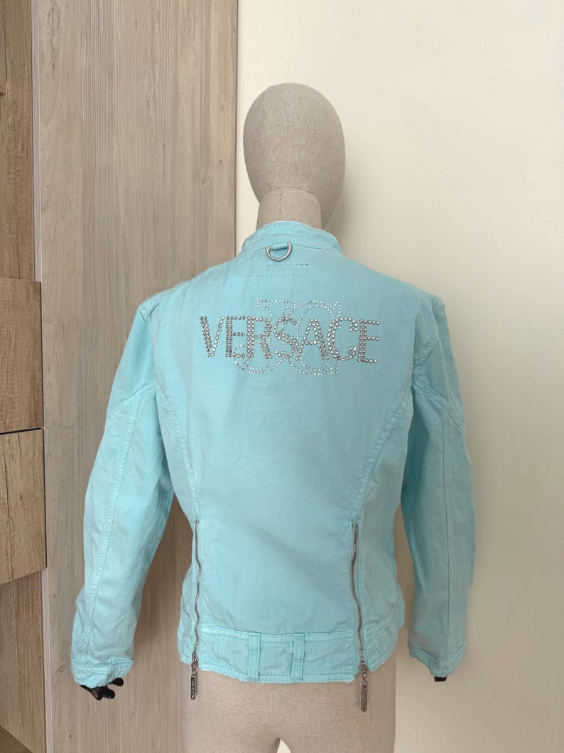 Może przedstawiać: Jasnoniebieska kurtka z napisem "VERSACE" wysadzanym srebrnymi kryształkami na plecach. Kurtka posiada zapięcie na zamek błyskawiczny i mały srebrny pierścień u g&oacute;ry plec&oacute;w. Kurtka jest prezentowana na manekinie.