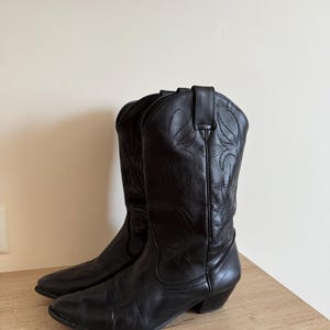 Peut inclure: Une paire de bottes de cowboy en cuir noir avec un bout pointu, un talon bottier et des motifs de surpiqûres ton sur ton sur la tige. Les bottes sont dotées de tirettes sur le dessus et d'une finition lisse, présentant une silhouette classique.