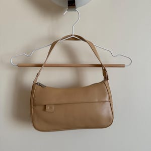 May include: A tan handbag with a silver zipper and a small square pocket is hanging on a white hanger with a wooden bar. The bag has a curved shape and a shoulder strap. The background is a plain, light-colored wall.