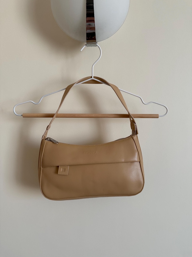 May include: A tan handbag with a silver zipper and a small square pocket is hanging on a white hanger with a wooden bar. The bag has a curved shape and a shoulder strap. The background is a plain, light-colored wall.