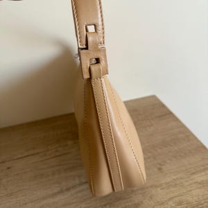May include: A tan leather shoulder bag with a curved shape. The bag features a matching handle with a rectangular buckle. The stitching is visible along the seams, and the bag is held by a hand.