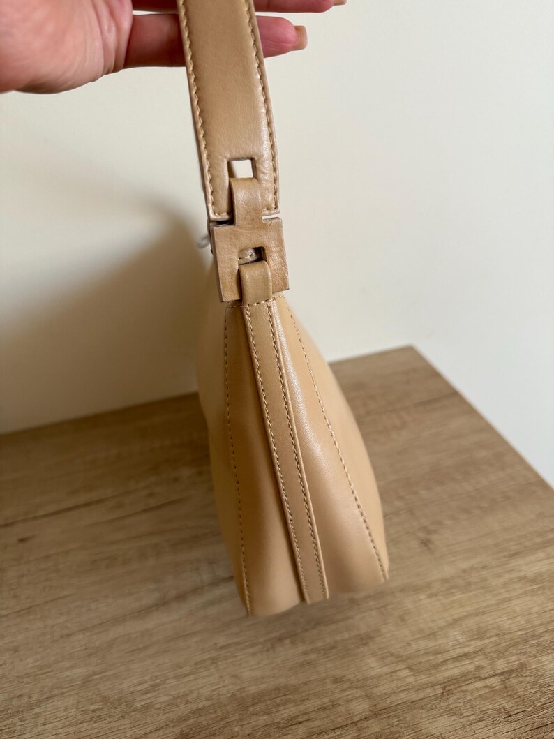May include: A tan leather shoulder bag with a curved shape. The bag features a matching handle with a rectangular buckle. The stitching is visible along the seams, and the bag is held by a hand.