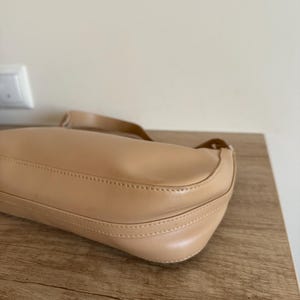 May include: A beige shoulder bag with a curved design. The bag is made of a smooth material and features a long strap. The bag is sitting on a wooden surface.
