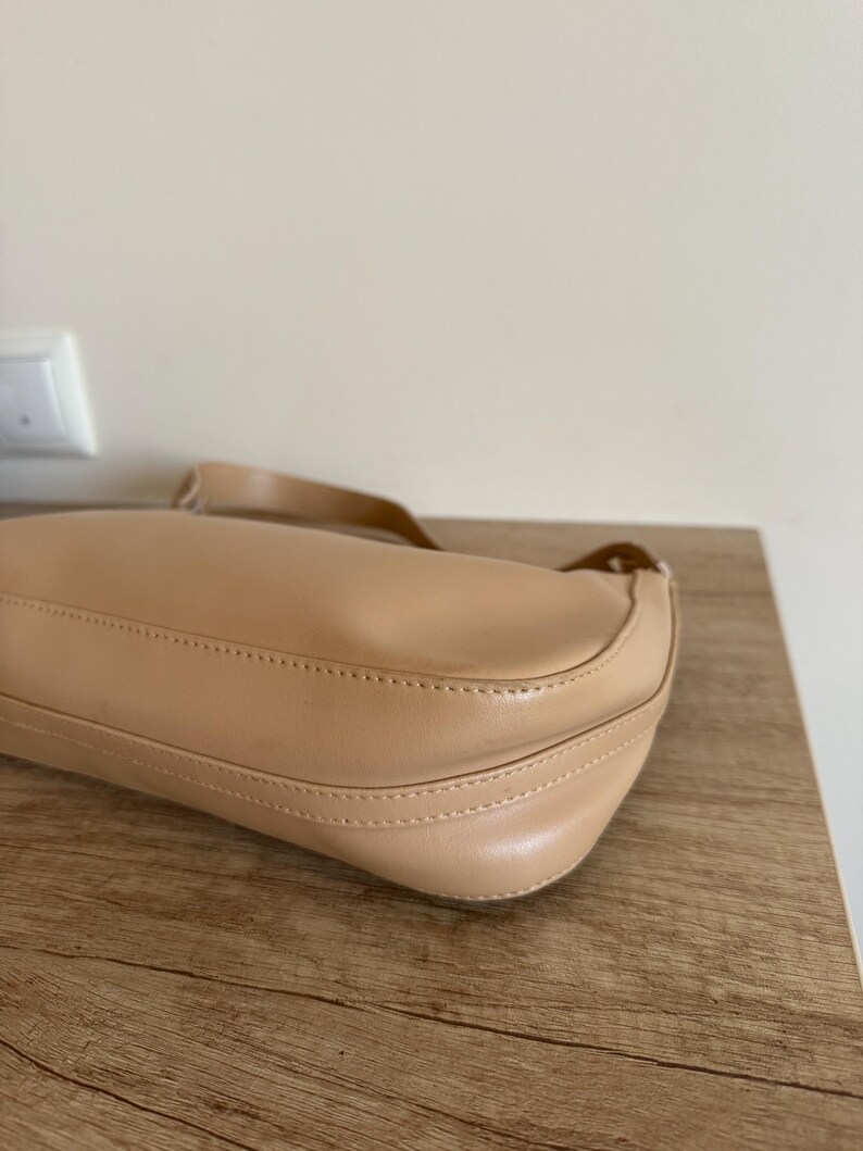 May include: A beige shoulder bag with a curved design. The bag is made of a smooth material and features a long strap. The bag is sitting on a wooden surface.