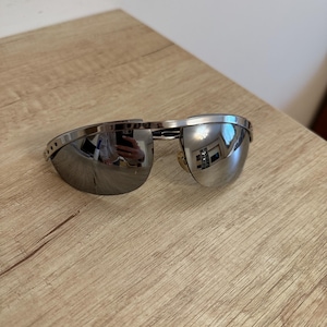 May include: A pair of sunglasses with a metallic silver frame and reflective, mirrored lenses. The design features a single continuous brow bar connecting the two lenses, with small circular ventilation holes visible on the side of the frame.