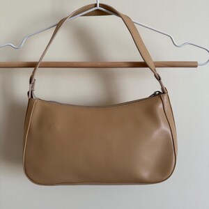 May include: A tan-colored shoulder bag with a rectangular shape and a curved top. The bag has a zipper closure and a single shoulder strap. The bag is hanging on a white hanger.