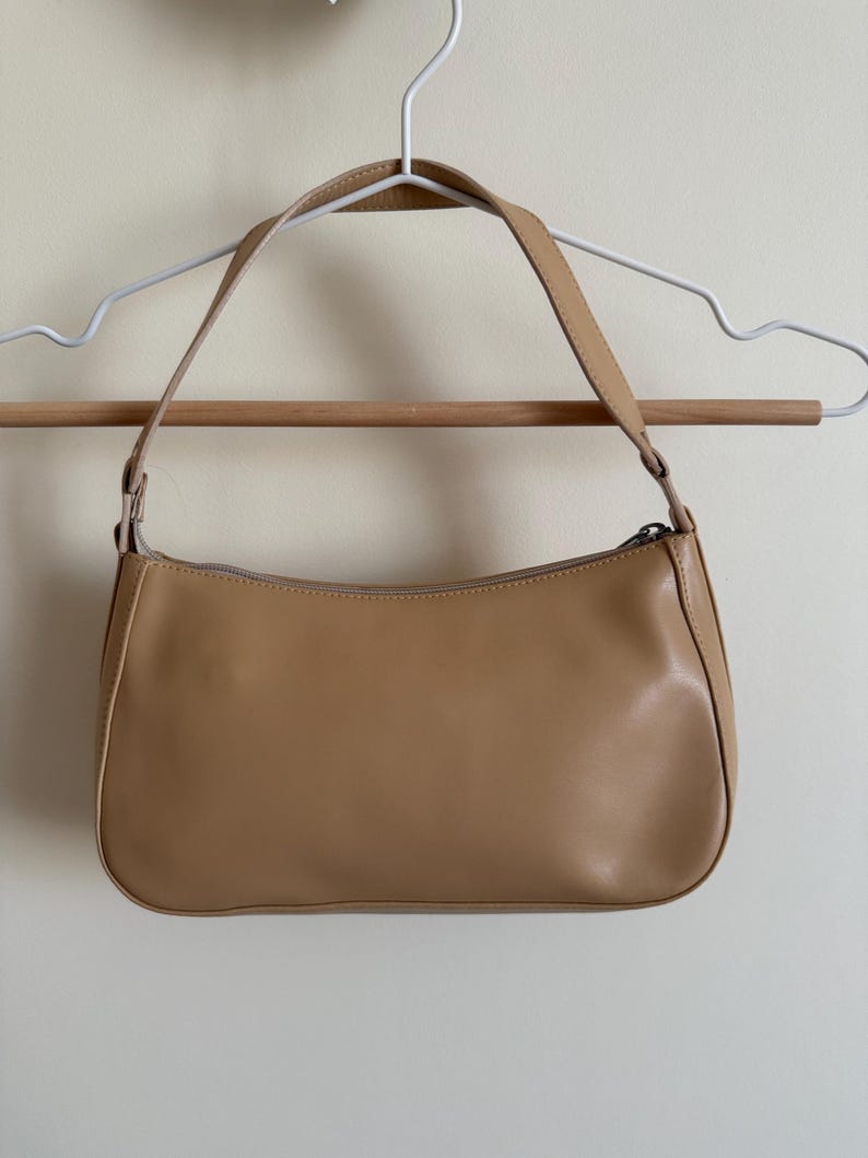 May include: A tan-colored shoulder bag with a rectangular shape and a curved top. The bag has a zipper closure and a single shoulder strap. The bag is hanging on a white hanger.