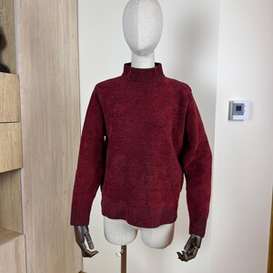 May include: A burgundy mock neck sweater is presented on a mannequin. The sweater features long sleeves and a ribbed texture. The mannequin has a neutral-toned head and torso, with black wooden hands.