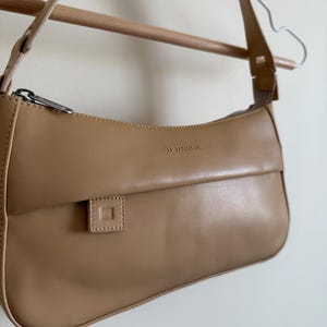 May include: A tan shoulder bag with a curved design, featuring a flap closure and a small square detail. The bag has a silver zipper and a long strap. The text "LE TANNEUR" is visible on the bag.