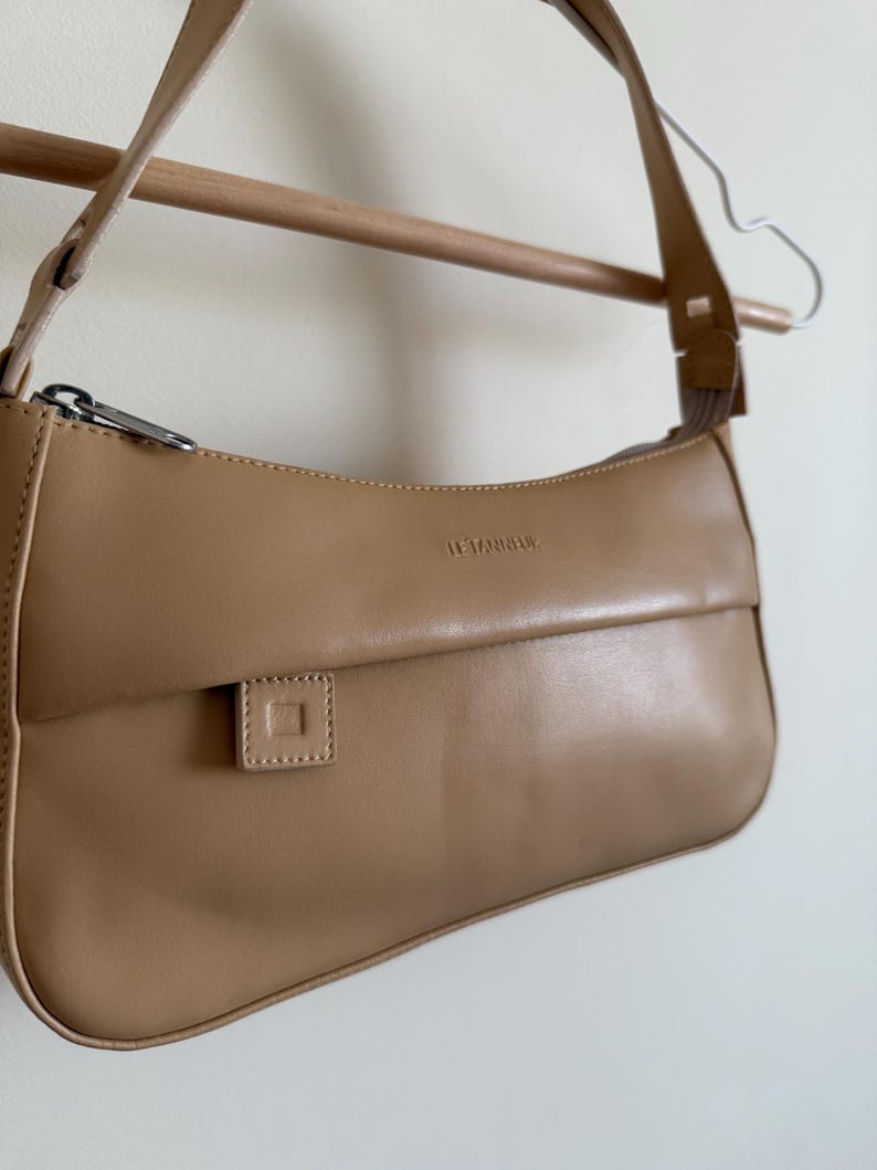 May include: A tan shoulder bag with a curved design, featuring a flap closure and a small square detail. The bag has a silver zipper and a long strap. The text "LE TANNEUR" is visible on the bag.