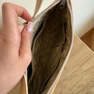 May include: Tan shoulder bag with a zipper closure. The interior features multiple pockets and a patterned lining. The bag has a long strap and a silver-toned zipper pull.