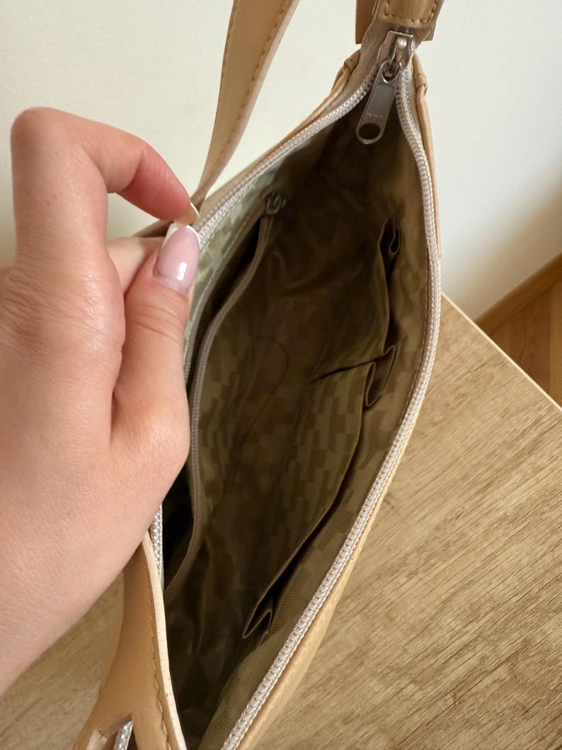 May include: Tan shoulder bag with a zipper closure. The interior features multiple pockets and a patterned lining. The bag has a long strap and a silver-toned zipper pull.
