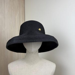 Puede incluir: Un sombrero negro para el sol con ala ancha y un pequeño botón dorado decorativo. El sombrero está hecho de material tejido y se exhibe en un maniquí. El ala del sombrero tiene aproximadamente 38 cm de diámetro.