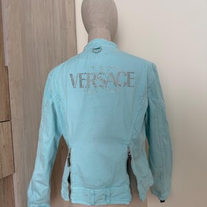 Może przedstawiać: Jasnoniebieska kurtka z napisem "VERSACE" wysadzanym kryształkami na plecach. Kurtka ma długie rękawy, st&oacute;jkę i dwa pionowe zamki z przodu. Kurtka jest prezentowana na manekinie.