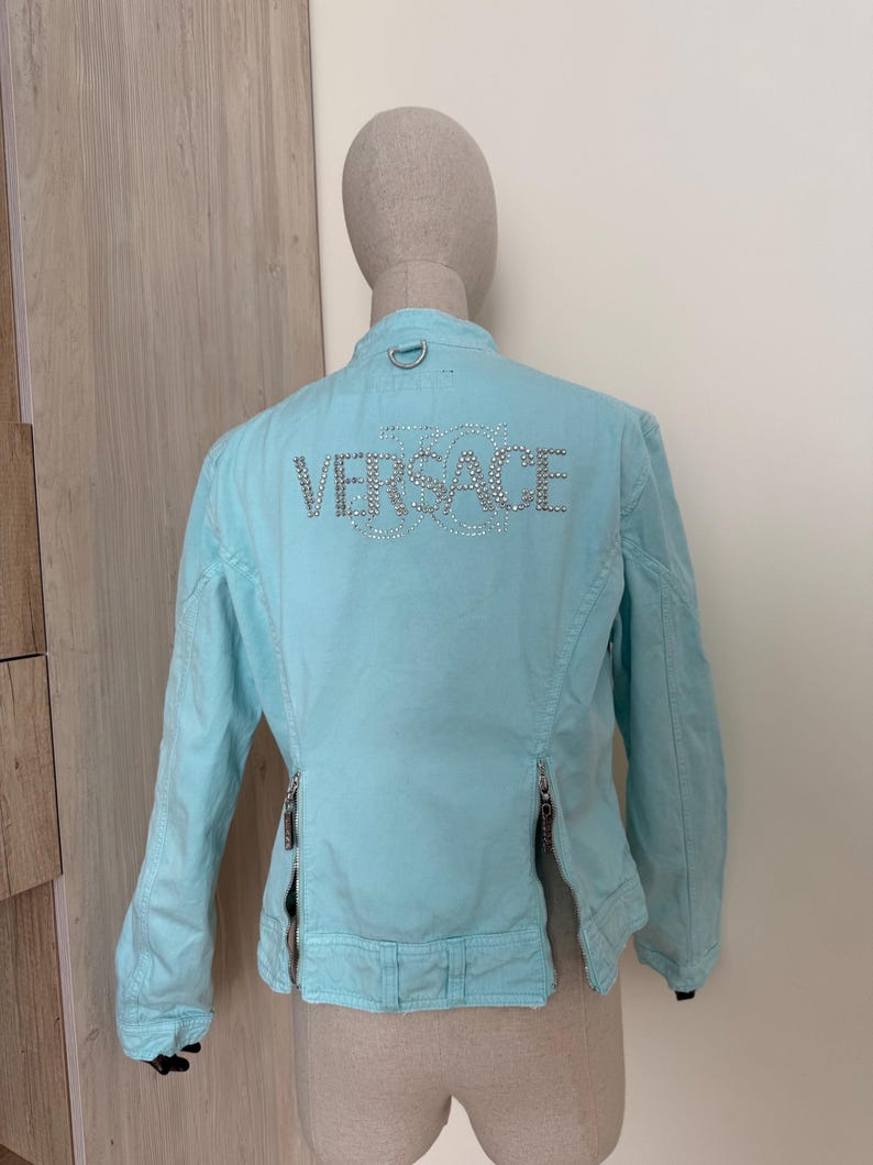 Może przedstawiać: Jasnoniebieska kurtka z napisem "VERSACE" wysadzanym kryształkami na plecach. Kurtka ma długie rękawy, st&oacute;jkę i dwa pionowe zamki z przodu. Kurtka jest prezentowana na manekinie.