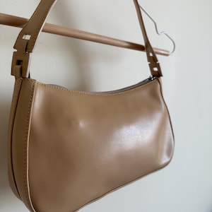 May include: A tan-colored shoulder bag with a curved shape and a long strap. The bag is made of a smooth material and features a zipper closure. The bag is hanging on a wooden rod and a metal hanger.