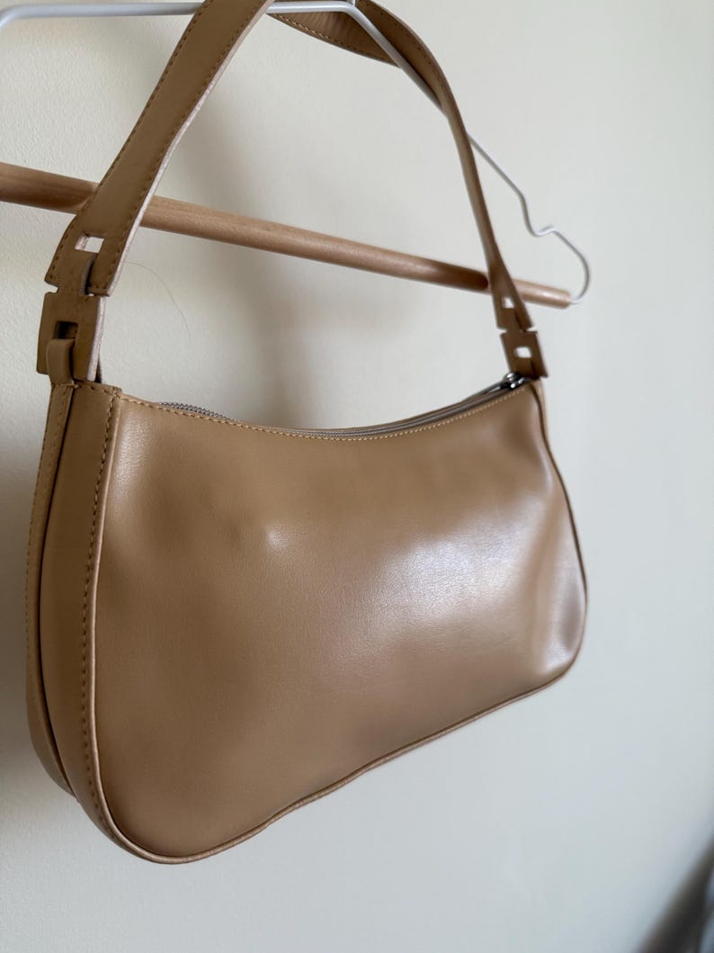 May include: A tan-colored shoulder bag with a curved shape and a long strap. The bag is made of a smooth material and features a zipper closure. The bag is hanging on a wooden rod and a metal hanger.