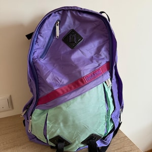 Può includere: Uno zaino vintage viola, verde acqua e rosa. Lo zaino ha più scomparti con cerniera, spallacci regolabili e un emblema nero. La parola "OPTIMAX" è stampata su una cinghia rosa. La borsa è in nylon.