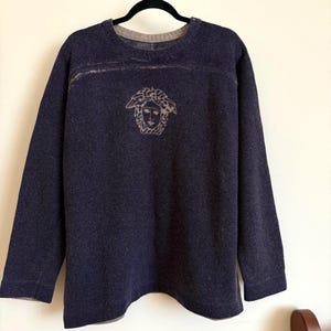 Puede incluir: Un suéter azul marino con cuello redondo y mangas largas. El suéter presenta un emblema de cabeza de Medusa en el centro y una banda de color más claro en el pecho. El suéter está colgado de una percha negra.