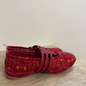 May include: A pair of red and gold patterned shoes with a strap closure. The shoes have a glossy finish and a rubber sole. The design features a floral pattern. The shoes are on a wooden surface.