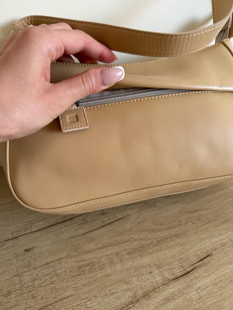 May include: A tan shoulder bag with a rectangular shape. The bag has a zipper closure and a matching strap. The bag is made of a smooth material and has a minimalist design.