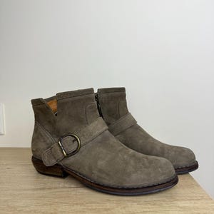 May include: A pair of gray suede ankle boots with a buckle detail. The boots have a low heel and a side zipper. The boots are displayed on a wooden surface.