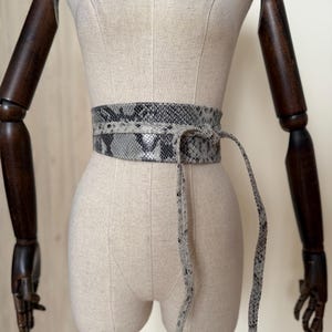 Puede incluir: Un cinturón ancho estilo obi con un patrón de piel de serpiente en gris y negro. El cinturón se envuelve alrededor de la cintura y se asegura con dos tiras largas, finas y a juego que cuelgan de un nudo frontal, creando un aspecto texturizado.