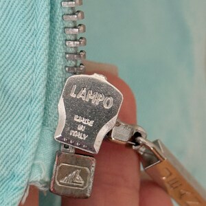 Może przedstawiać: Zbliżenie srebrnego metalowego zamka błyskawicznego z nazwą marki "LAMPO" i napisem "MADE IN ITALY" wytłoczonym na suwaku. Zamek błyskawiczny jest przymocowany do jasnoniebieskiej tkaniny.