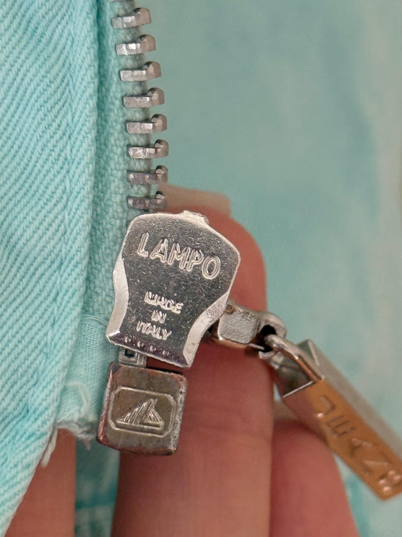 Może przedstawiać: Zbliżenie srebrnego metalowego zamka błyskawicznego z nazwą marki "LAMPO" i napisem "MADE IN ITALY" wytłoczonym na suwaku. Zamek błyskawiczny jest przymocowany do jasnoniebieskiej tkaniny.