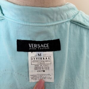 Może przedstawiać: Zbliżenie metki koszuli Versace Jeans Couture w kolorze jasnoniebieskim. Czarna metka zawiera nazwę marki, rozmiar M. Metka zawiera r&oacute;wnież napis "MADE IN ITALY" i inne szczeg&oacute;ły.