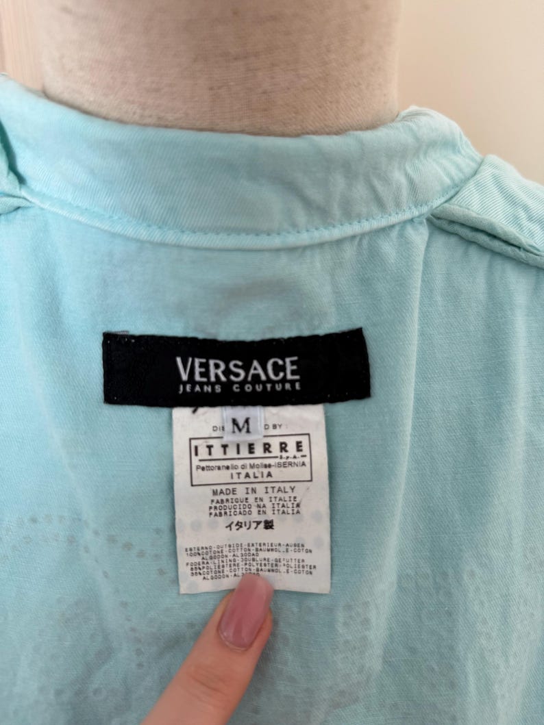 Może przedstawiać: Zbliżenie metki koszuli Versace Jeans Couture w kolorze jasnoniebieskim. Czarna metka zawiera nazwę marki, rozmiar M. Metka zawiera r&oacute;wnież napis "MADE IN ITALY" i inne szczeg&oacute;ły.