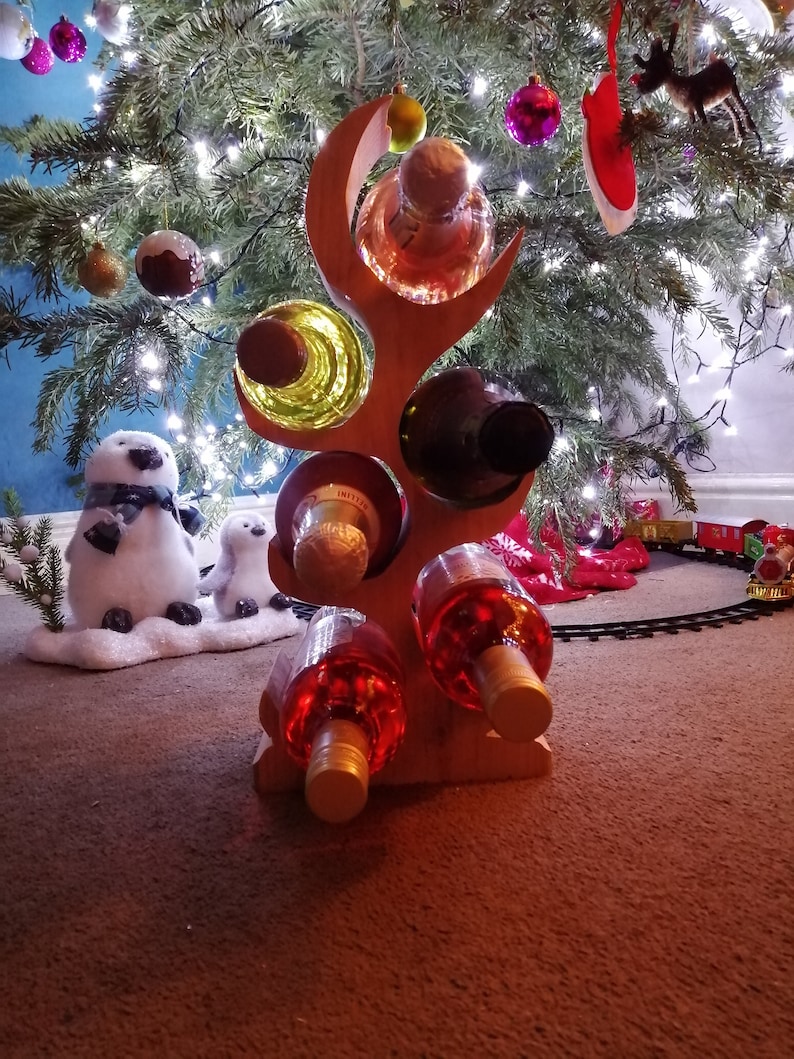 May include: Wooden wine rack shaped like a tree branch with five bottles of wine. The rack is decorated with a red and white Christmas ornament and is sitting under a Christmas tree.