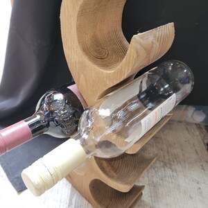 May include: Wooden wine bottle holder with a natural wood finish. The holder has three slots for bottles and is shaped like a stylized tree. Two bottles of wine are in the holder, one red and one white.