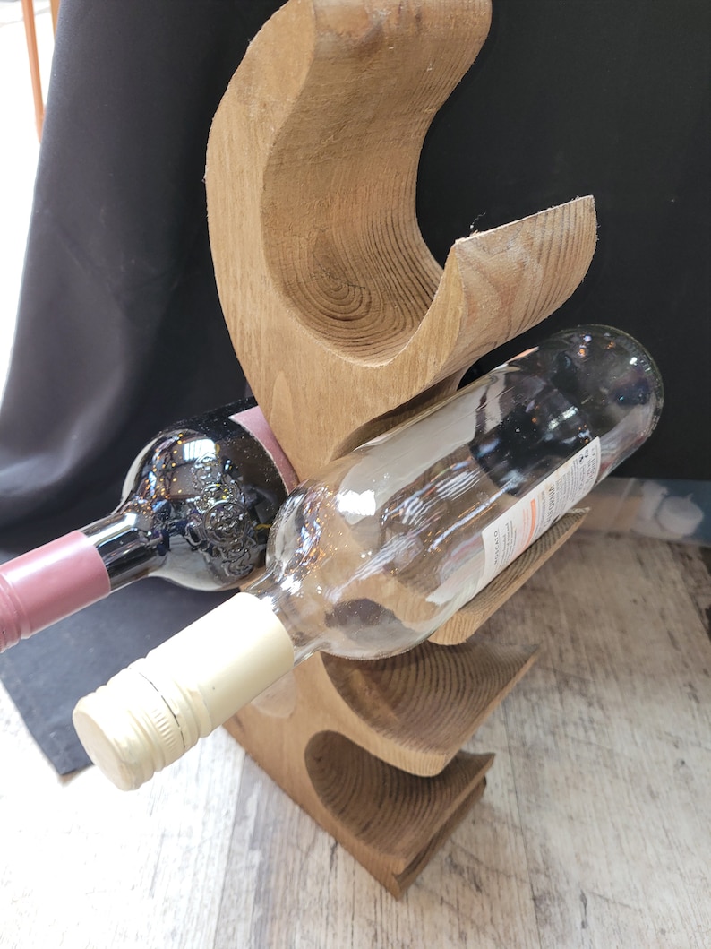 May include: Wooden wine bottle holder with a natural wood finish. The holder has three slots for bottles and is shaped like a stylized tree. Two bottles of wine are in the holder, one red and one white.