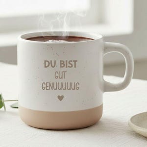 Könnte beinhalten: Ein weißer Keramikbecher mit gesprenkelter Oberfläche und beigem Boden. Der Becher trägt den braunen Schriftzug DU. BIST. GUT GENUUUUUG und ein kleines Herzsymbol. Dampf steigt aus der dunklen Flüssigkeit im Becher auf, der einen Henkel besitzt.