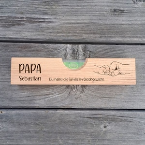 Könnte beinhalten: Eine Holz-Wasserwaage mit dem Text "PAPA Sebastian" und einer Faust-Stoß-Grafik. Die Wasserwaage hat eine klare Ampulle mit einer grünen Flüssigkeitsblase. Der Text "Du hältst die Familie im Gleichgewicht" ist ebenfalls vorhanden.