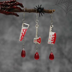 May include: Silver-toned Halloween earrings featuring a saw, axe, and cleaver design with red accents, each dangling a red teardrop bead. A black spider sits on a branch above the earrings, with a spiderweb in the background.