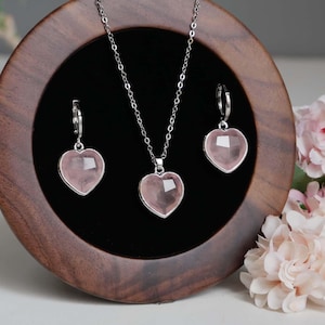May include: A jewelry set featuring a silver-toned chain necklace and two matching hoop earrings, each adorned with a faceted, heart-shaped light pink stone pendant. The pieces are displayed on a circular dark wood stand with a black velvet center.