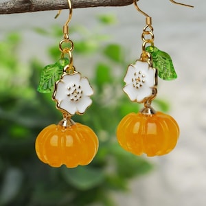 Peut inclure: Une paire de boucles d'oreilles pendantes dorées avec une fleur blanche et un petit charme de feuille verte, et un charme de citrouille orange vif.