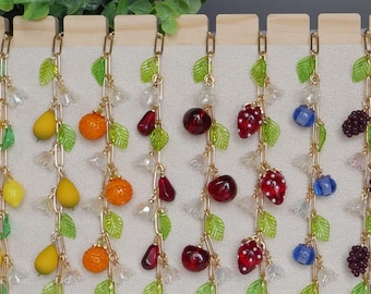 May include: A collection of gold-tone metal charm bracelets displayed on a stand. Each bracelet features a unique fruit or berry motif, including grapes, lemons, pears, oranges, cherries, strawberries, blueberries, blackberries, and pomegranates.