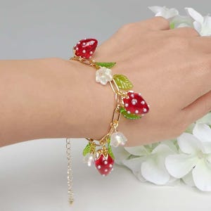 Peut inclure: Un bracelet en or avec des breloques en forme de fraises rouges et blanches et des feuilles vertes. Le bracelet est porté au poignet.