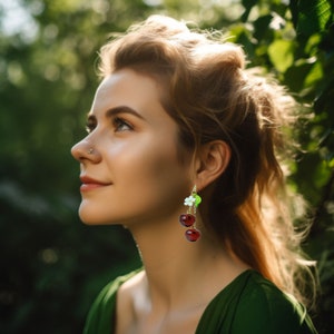 May include: A woman with long, wavy brown hair wearing a green dress and cherry earrings. The earrings are made of red glass beads and have a small green leaf charm.