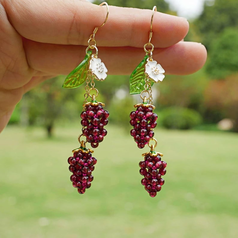 Op de afbeelding: Een paar oorhangers met drie trossen donkerrode kralen, elke tros lijkt op een tros druiven. De oorhangers zijn versierd met groene bladeren en witte bloemen.