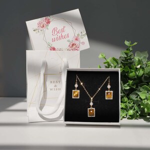 May include: A white gift box with a gold and white patterned design. The box contains a gold necklace and earrings with a square-shaped pendant and a tiger's eye stone. The box is sitting on a white surface with a white gift bag and a card that says "Best wishes".