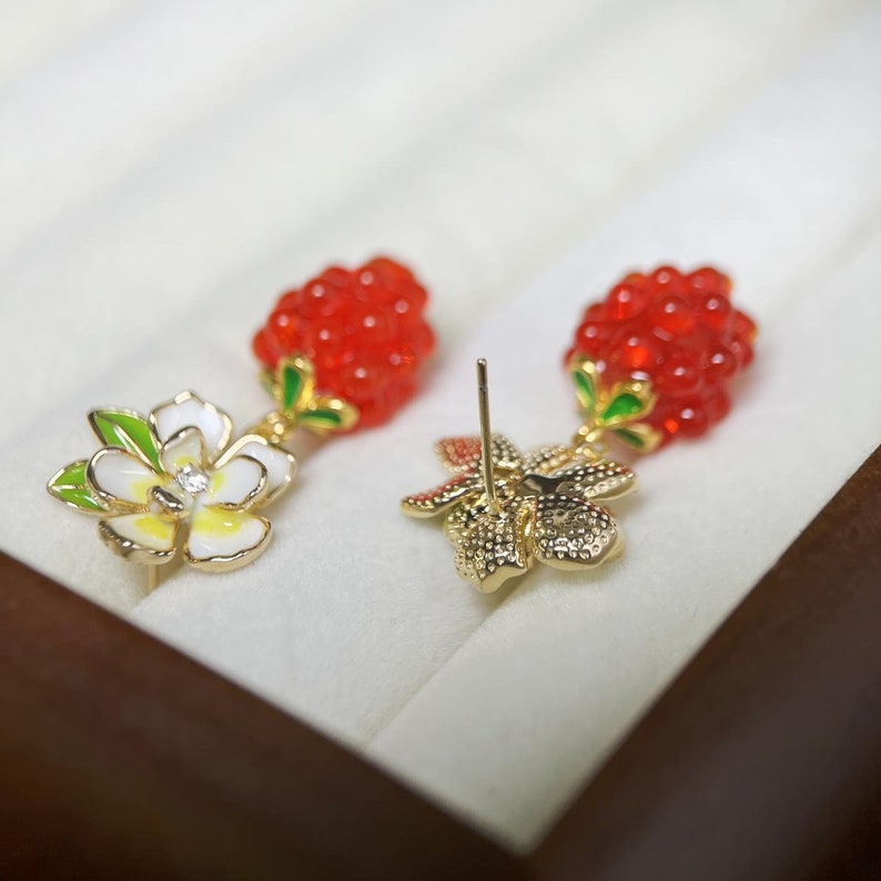 May include: A pair of dangle earrings with a red berry cluster and a white flower with a green stem. The earrings have gold-tone metal findings.