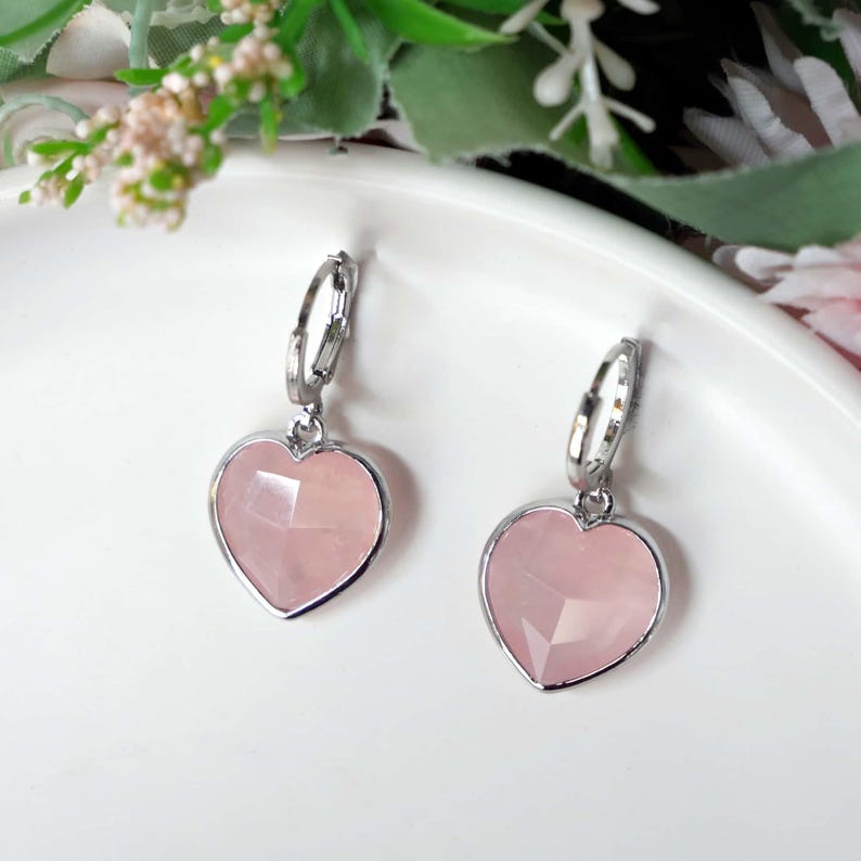 May include: A pair of silver-toned hoop earrings featuring faceted, heart-shaped light pink stone pendants. Each earring consists of a small circular hoop connected to a heart-shaped charm with a smooth, polished silver-colored metal border.