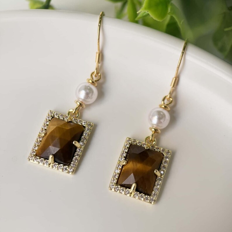 May include: Gold earrings with a brown tiger's eye stone and a white pearl dangle. The stone is set in a square frame with clear rhinestones.