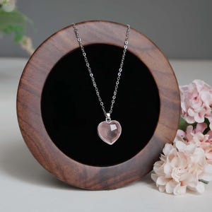 May include: A silver-toned chain necklace featuring a faceted, heart-shaped light pink stone pendant. The necklace is displayed on a circular dark wood stand with a black velvet interior, positioned next to light pink floral accents.