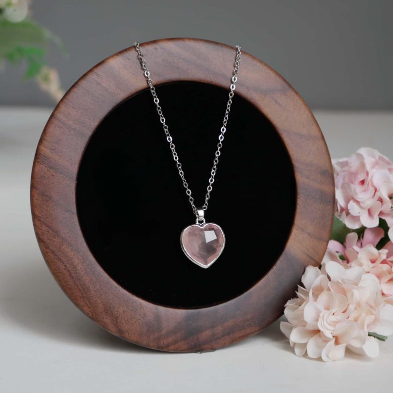 May include: A silver-toned chain necklace featuring a faceted, heart-shaped light pink stone pendant. The necklace is displayed on a circular dark wood stand with a black velvet interior, positioned next to light pink floral accents.